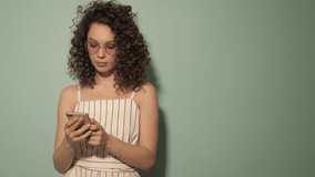 Raw video.Beautiful smiling model with afro curls hairstyle dressed in summer hipster clothes.Carefree girl posing in studio.Positive woman chatting with friends.She using smartphone.Texting sms