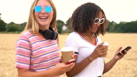 Slow motion tracking video clip of pretty blonde girl and mixed race teenager young women wearing sunglasses drinking coffee and using mobile cell smart phones for social media