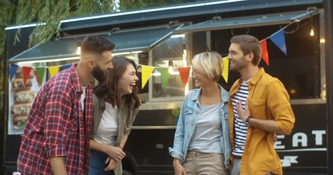 Two young happy Caucasian couples standing outdoor at festive food truck cafe and talking cheerfully. Stylish joyful men and women in hugs laughing and chatting loudly while joking at outside bar.
