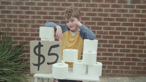 Little boy has a toilet paper stand outside of his house selling rolls of toilet paper taking advantage of a toilet paper shortage. There is a hand painted sign with five dollars written on it.