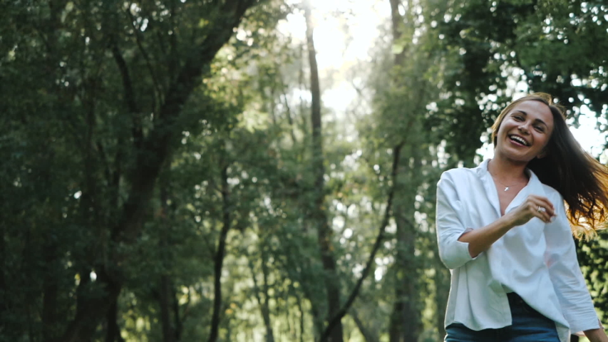 Front view of a young pretty girl in a white shirt and long hair who is looking at the camera and bouncing for a nice fun shot in a warm cozy park among green trees and the shining sun between them.