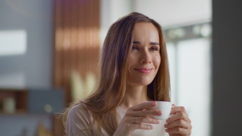 Portrait of satisfied woman drinking tea on kitchen. Attractive lady enjoying cup of coffee at home in slow motion. Smiling woman holding cup of tea in hands at morning