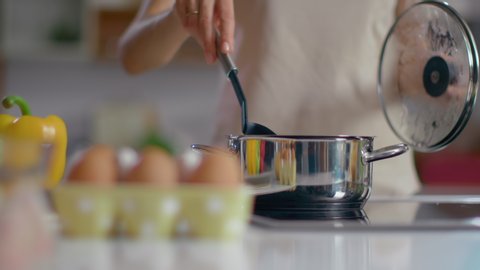Unrecognizable woman cooking soup on kitchen. Close up of housewife stirring soup with spoon at home in slow motion. Woman chef standing at stove on domestic kitchen