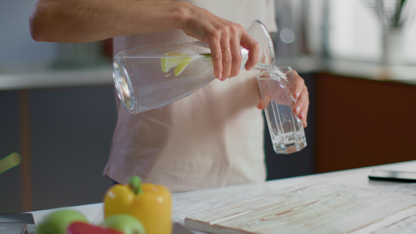 Close up of woman pouring fresh water into glass from jug at home. Attractive girl drinking water on domestic kitchen. Young woman holding glass of water with lemon on kitchen in slow motion Royalty-Free Stock Footage #1047905347