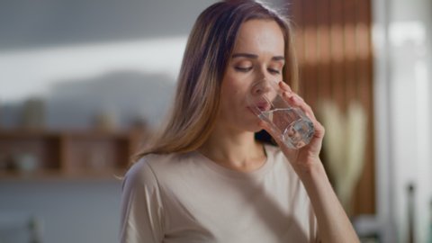 Close up of woman pouring fresh water into glass from jug at home. Attractive girl drinking water on domestic kitchen. Young woman holding glass of water with lemon on kitchen in slow motion