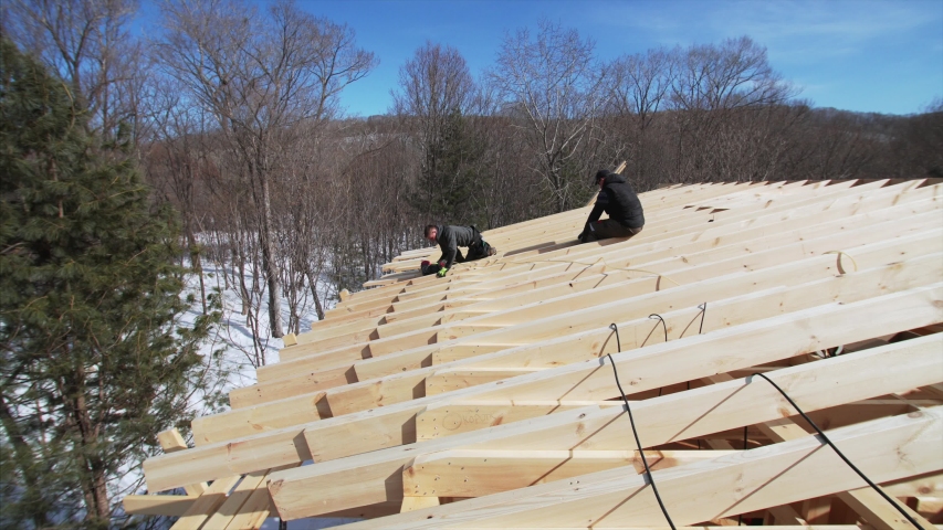 Sliding view of builders nailing wooden planks on the roof with pneumatic nail gun. Frame house under construction. Sunny winter day. Leafless forest is on the background.