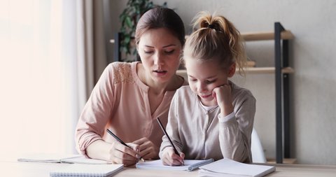 Cute small 6-7 years kid daughter learning writing with young mom tutor. Adult parent mother teaching school child girl helps with homework studying sitting at home table. Children education concept