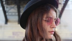 4K video cute stylish young girl in pink glasses a light dress and a black hat on a background of a round dark bridge with hair in the wind