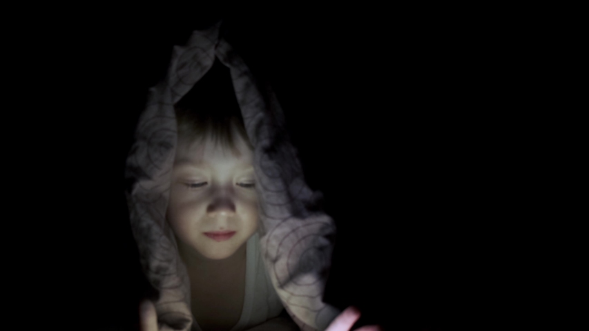 little boy plays on tablet while lying under blanket. watching cartoons hiding.