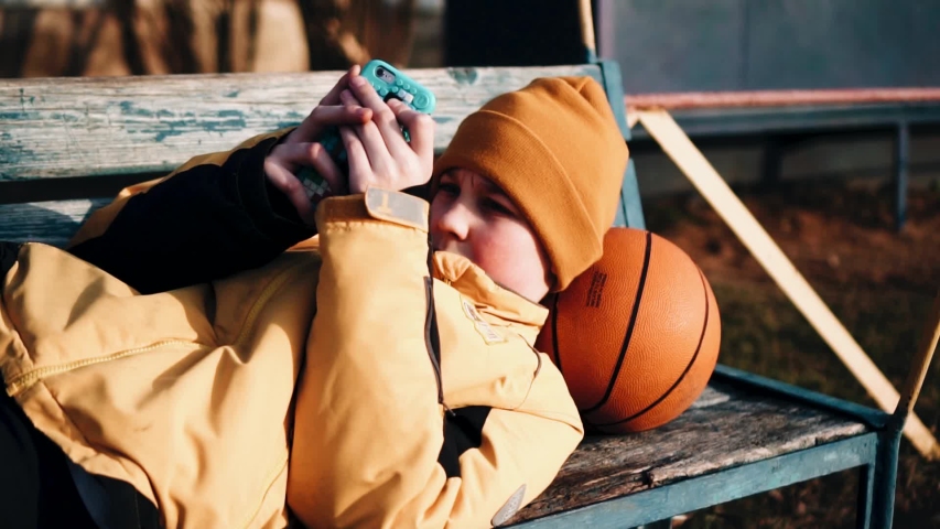 The boy put a ball under his head and lies on the bench. Quiet rest after the game. Beautiful light from the evening sun.