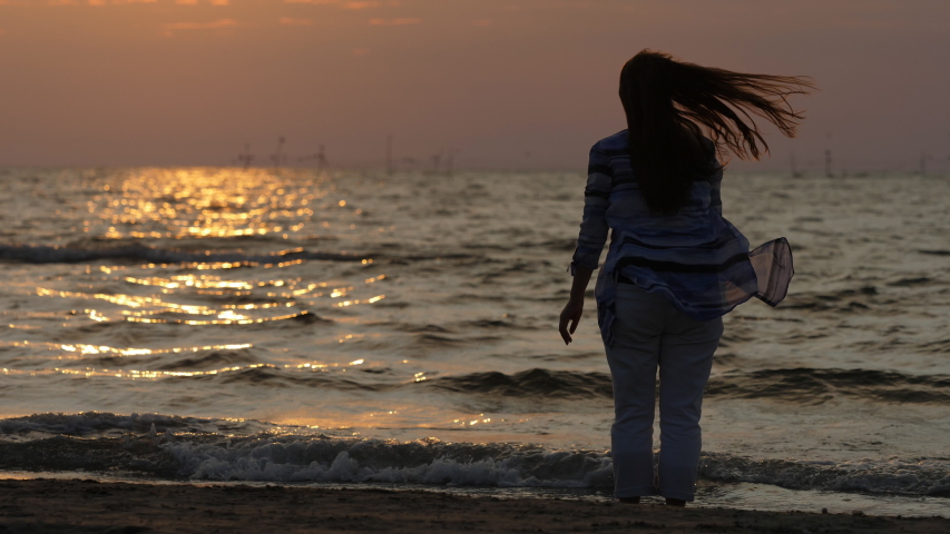 Single woman silhouette stand at sea shore and look at sunrise, long hair move , slow motion