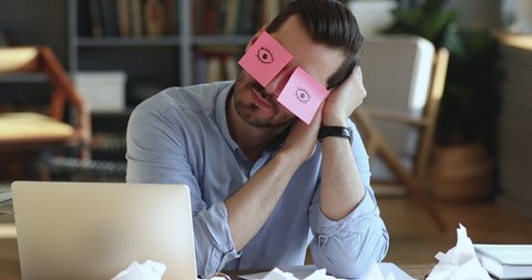 Funny lazy office worker napping at workplace covering eyes with sticky notes. Inefficient tired male employee pretends working sleeping with stickers on face sits at desk. Cheating to sleep concept