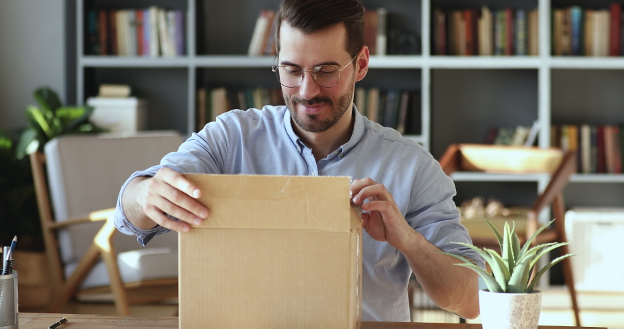 Smiling young man customer opening parcel cardboard box sitting at home office desk. Happy consumer unpacking postal shipping delivery satisfied with good purchase. Fast post shipment service concept | Shutterstock HD Video #1047963097