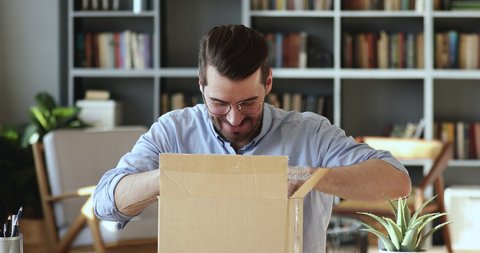 Smiling young man customer opening parcel cardboard box sitting at home office desk. Happy consumer unpacking postal shipping delivery satisfied with good purchase. Fast post shipment service concept