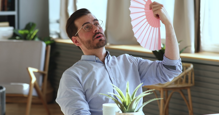 Exhausted young man waving fan suffering from summer heat inside apartment. Sweaty overheated businessman feeling hot uncomfortable frustrated about humidity, broken air conditioner in office concept. Royalty-Free Stock Footage #1047963109