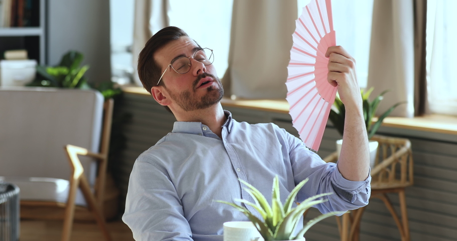 Exhausted young man waving fan suffering from summer heat inside apartment. Sweaty overheated businessman feeling hot uncomfortable frustrated about humidity, broken air conditioner in office concept. | Shutterstock HD Video #1047963109
