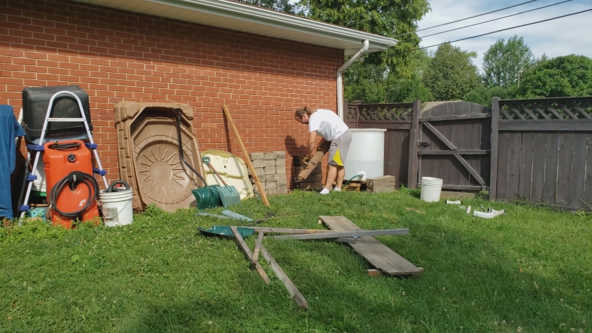 Man with glasses and ponytail wearing white t-shirt and shorts working in back yard next to brick wall cleaning or organizing some stuff or items.