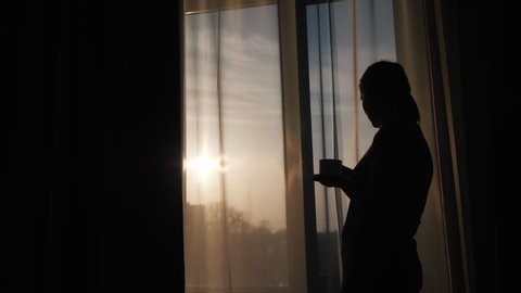 The silhouette of a woman at the window, drinks a hot drink and looks at the rising sun