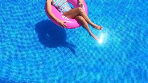 Top view of a young woman with sunglasses, hat and swimsuit in a blue pool. Pretty girl on a pink float enjoying the summer.