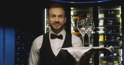 Portrait of Caucasian handsome male waiter smiling to camera with tray and two empty glasses for wine. Close up of attractive man working in restaurant. Worker serving drinks.