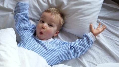 Cute funny cheerful little child boy in blue pajamas wakes up in the morning in bed, stretches and hides under the blanket again.