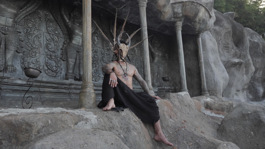 Tattooed masked skull ethnic pagan shaman sit on stage an ancient temple