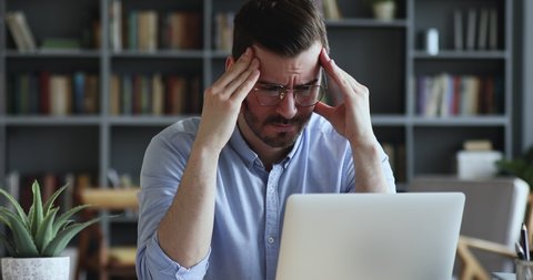 Stressed frustrated businessman wearing glasses suffering from headache at work. Exhausted tensed worker coping with migraine, emotional stress concept feeling pain sitting at office desk with laptop.