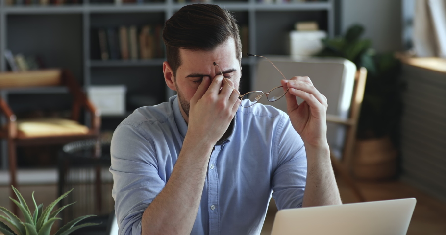 Exhausted young businessman professional taking off glasses massaging dry tired irritated eyes. Overworked worker feeling eye strain concept. Headache and fatigue after office work, computer syndrome. | Shutterstock HD Video #1047984337