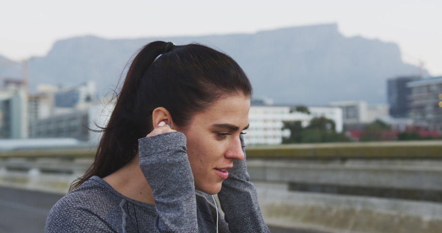 Side view close up of a fit Caucasian woman with long dark hair wearing sportswear, exercising outdoors in the city, listening to music on earphones, taking break standing in the street, putting on