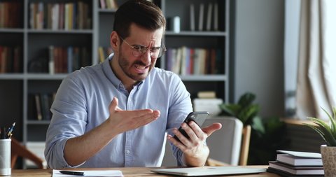 Mad unhappy businessman feeling annoyed using smart phone. Angry businessman having problem with mobile spam message. Male user frustrated by low battery or bad signal concept sitting at office desk.