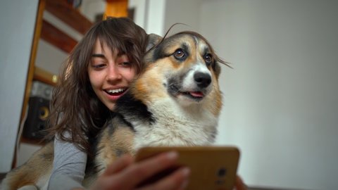joyful smiling attractive young woman hugging cute corgi dog and taking selfie with pet using phone