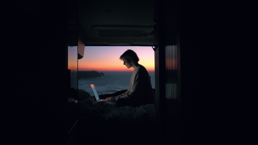 A young entrepreneur woman is working on her computer in a camper van. The door is open to an epic view of the sea in a sunset / sunrise. Remote working anywhere, the concept of digital nomad.