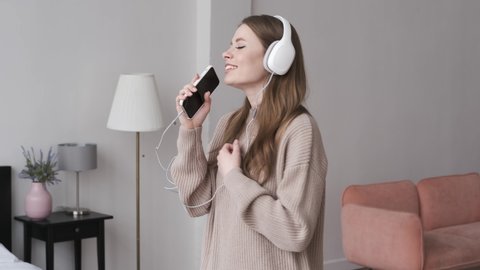 Joyful woman holding smart phone and singing karaoke. Young girl having fun alone at home. Happy girl in headphones laughing smiling and emotionally singing a song. Entertainment on weekend. Indoors