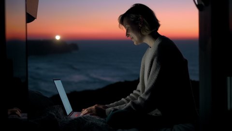 A young entrepreneur woman is working on her computer in a camper van. The door is open to an epic view of the sea in a sunset / sunrise. The lighthouse is in the background. Remote working anywhere. Stock Video