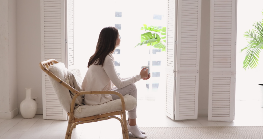 Relaxed young woman drinking tea looking through window enjoying peaceful lounge at home. Serene adult girl sitting in comfortable chair in cozy apartment dreaming, relaxing feeling no stress concept.