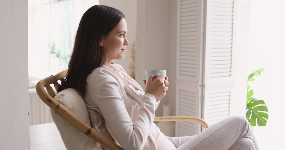 Serene carefree young woman drinking warm tea, breathing fresh air at home. Relaxed calm lady holding cup relaxing sitting in chair indoors. Smiling girl enjoys good morning feels stress free concept. Royalty-Free Stock Footage #1048023904