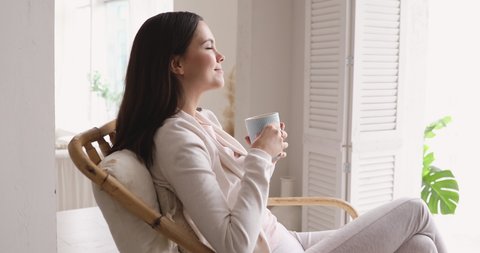 Serene carefree young woman drinking warm tea, breathing fresh air at home. Relaxed calm lady holding cup relaxing sitting in chair indoors. Smiling girl enjoys good morning feels stress free concept.