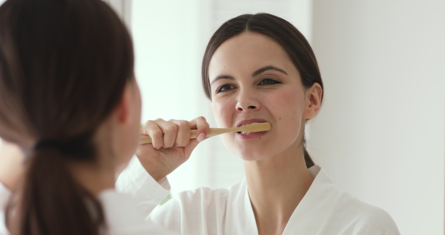 Happy young woman holding wooden toothbrush brushing teeth looking in mirror. Beautiful girl cleaning mouth doing morning dental care routine in bathroom. Lady using natural bamboo eco brush concept Royalty-Free Stock Footage #1048023928