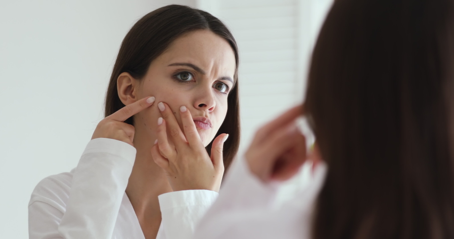 Upset teen girl feeling worried about facial skin problem looking in mirror. Depressed young woman touching face frustrated by blackheads, acne and pimples. Blemish skincare beauty treatment concept | Shutterstock HD Video #1048023994
