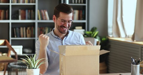 Excited man customer winner opening cardboard parcel box receiving gift sitting at home office desk. Overjoyed male consumer shopper unpacking postal delivery celebrating good online purchase concept.