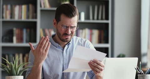 Angry stressed businessman opening envelope reading bad news in mail letter. Mad man feels frustrated about high bills, dismissal notice, bank debt, tax invoice or mistake sits at home office desk