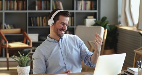 Funny happy young businessman wears wireless headphones having fun listening music. Smiling employee feels no stress at work pretending playing guitar. Cheerful man enjoys relaxing at home office desk