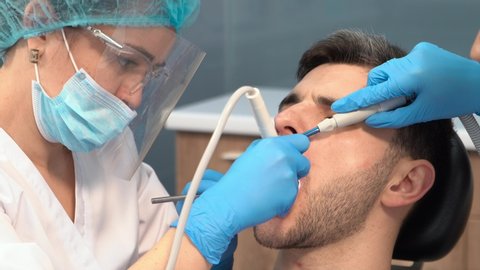 a professional dentist in a protective shield, goggles and face mask with the help of an assistant and modern equipment works with a male patient sitting with his mouth open. dental treatment. caries