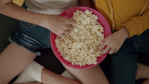 Dolly In Top View of the Hands of Two Teenage Girls Eating Pop Corn From a Pink Bowl Sitting on Couch While Watching a Movie Film. Concept of Low Fat Snacks and Good Sources of Dietary Fibers.