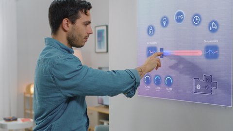 Handsome Young Man Interacts with Augmented Reality Display. He is Changing Climate Control Setting to a Higher Temperature. Smart Home Concept with Transparent Screen Made with VFX Special Effects.