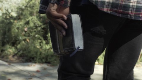 Man walking away from camera on the side walk holding the bible by his side.