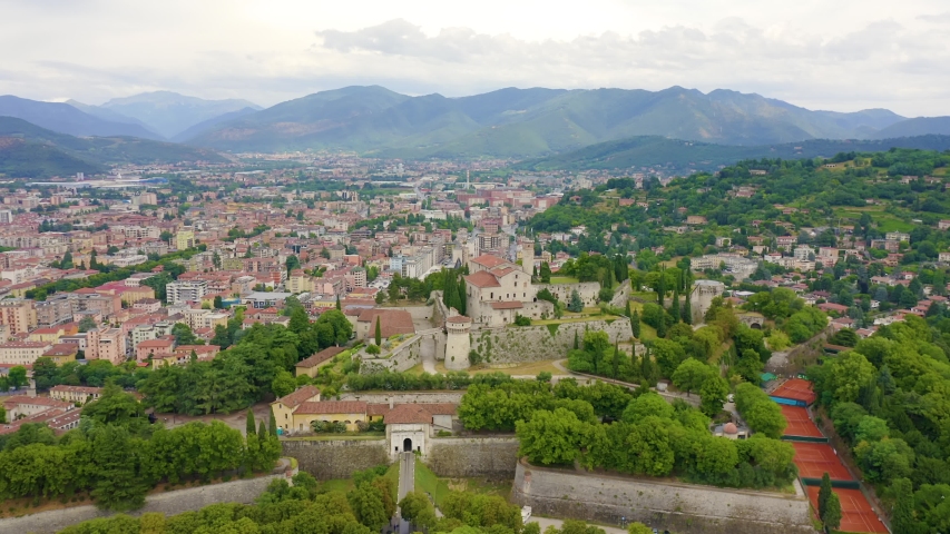 Brescia, Italy. Castello di Brescia, a medieval hilltop fortress perched on Cidneo Hill, dominates the city skyline with its stone walls, bastions, and historic towers surrounded by dense greenery. The aerial footage captures a slow orbital flight around the fortified complex, revealing the sprawling urban landscape of Brescia below with its characteristic red-tiled rooftops, while the forested slopes of the Prealps rise in the background under an overcast summer sky. The castle complex, one of the largest fortified structures in Europe, shows multiple layers of defensive walls, a central keep, and rounded towers integrated into the wooded hilltop. The surrounding city unfolds in all directions with a mix of historic and modern buildings, broad avenues, and patches of green parkland, offering a sweeping panoramic view from the elevated vantage point. The muted daylight and soft cloud cover create a calm, atmospheric mood over the entire scene, Point of interest