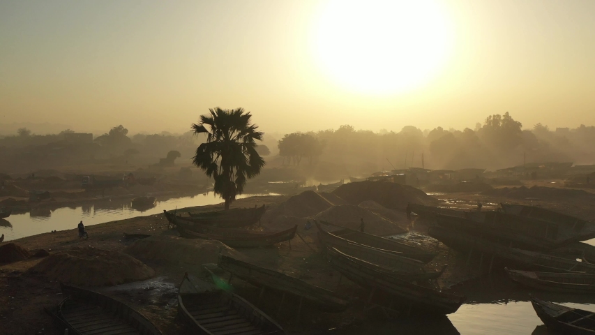 The drone flies looking into the sun towards and over a local brown sandy harbour with many wooden boats and local people with the sunrays seeping through the trees in Mali Aerial Drone Footage 4K