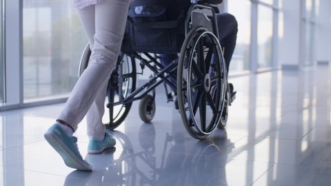 Low-section shotof nurse pushing driving wheelchair with handicapped patient through the corridor with mirror floor