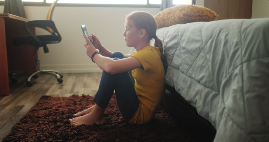 Text message on mobile telephone for bullying a sad little girl in bedroom at home. Young people looking at cell phone. Redhead female child with emotions, feelings of sadness and loneliness Royalty-Free Stock Footage #1048117030